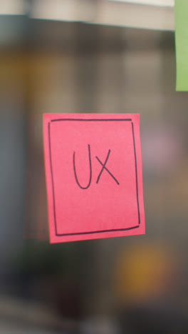 Vertical-Video-Close-Up-Of-Woman-Putting-Sticky-Note-With-UX-Written-On-It-Onto-Transparent-Screen-In-Office